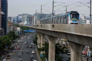 A Hyderabad Metro train traveling on an elevated corridor above a busy city road, representing ongoing discussions about the proposed metro takeover and its financial implications for Telangana.