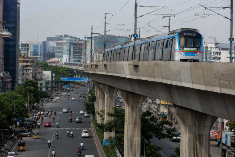 A Hyderabad Metro train traveling on an elevated corridor above a busy city road, representing ongoing discussions about the proposed metro takeover and its financial implications for Telangana.